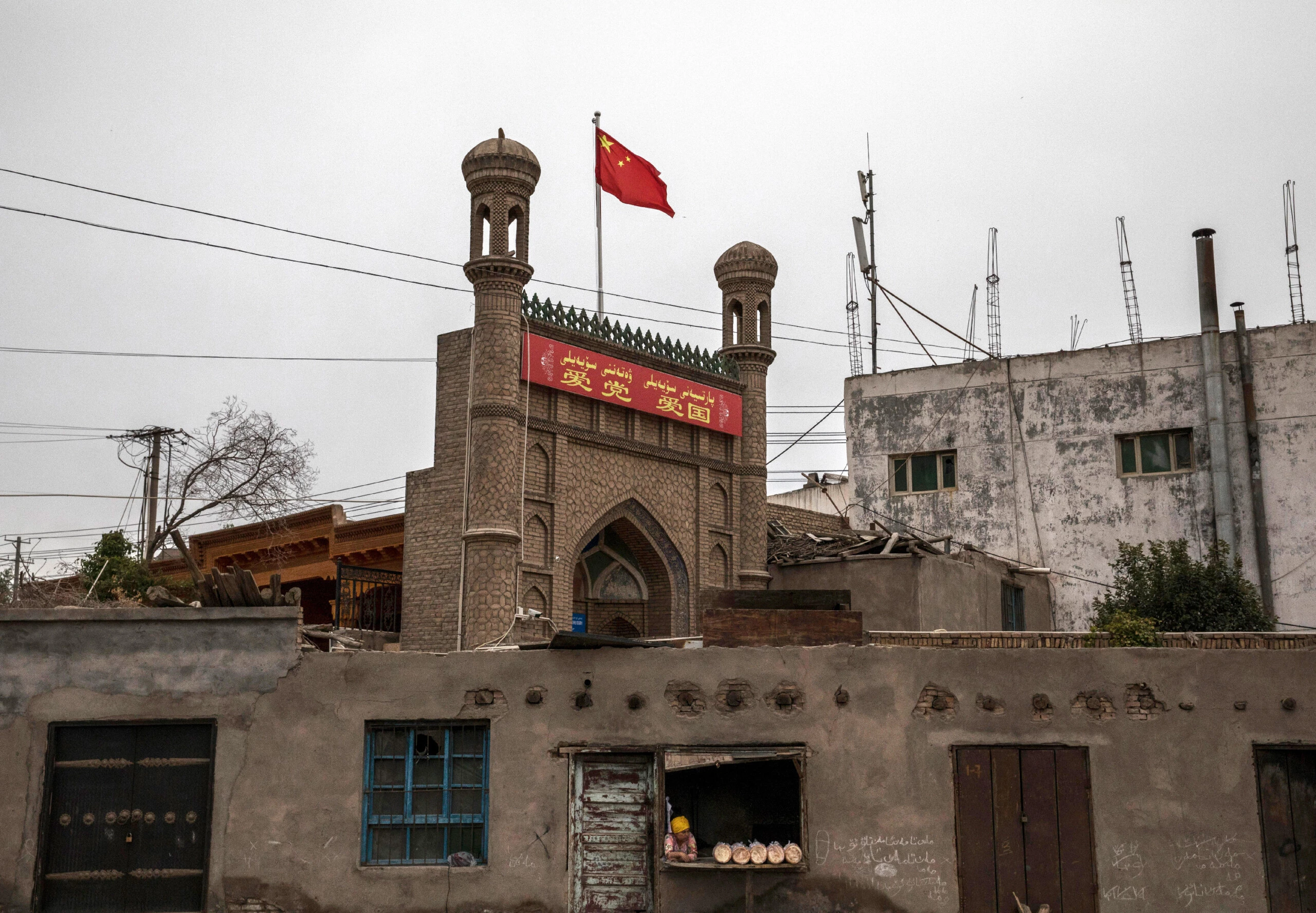 Uma bandeira chinesa tremula sobre uma mesquita local que havia sido fechada pelas autoridades, enquanto uma mulher de etnia uigur vende pão em sua padaria, em 28 de junho de 2017, na cidade antiga de Kashgar, no extremo oeste da província de Xingjiang, na China.