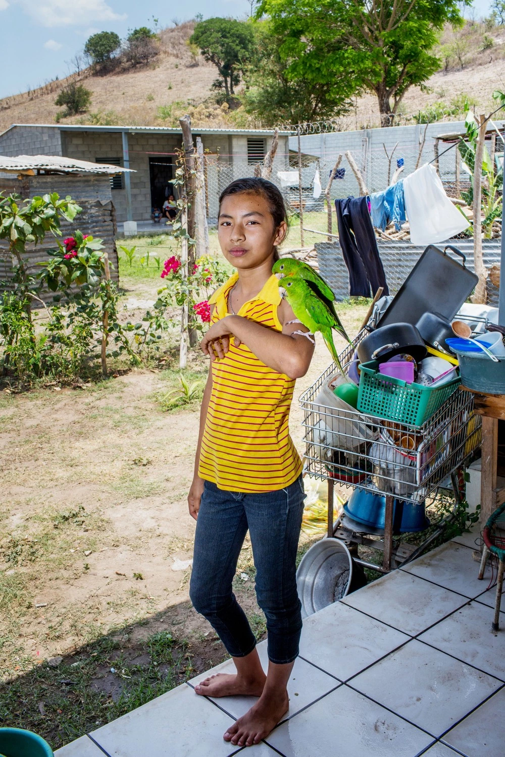 April 2017, El Salvador, San Salvador. 13 year old Kenya Fabiola Gonzales Trinidad holds her pet parrots on her arm at her home outside of San Salvador. She has just moved to the new community of new cinder block 3 room houses built with church donations, because her old neighborhood where she lived with her brother and grandfather was too dangerous due to gang activity. (Natalie Keyssar)