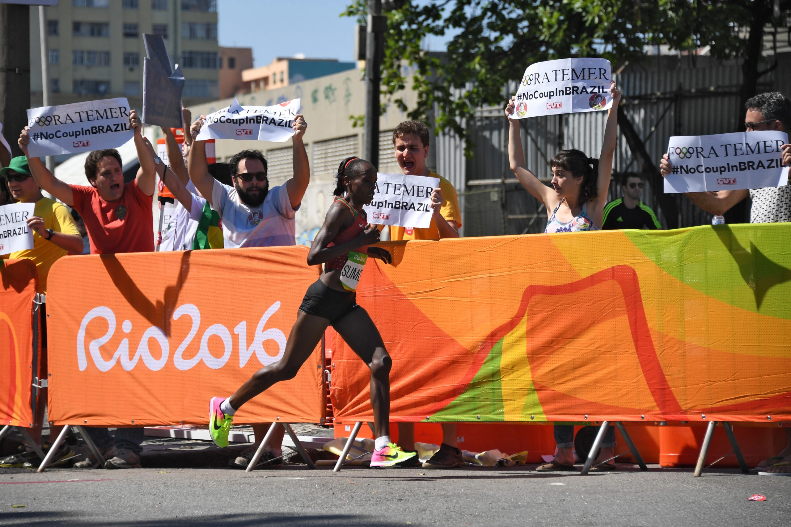 RIO DE JANEIRO, BRAZIL - AUGUST 14:  Kenya's Jemima Jelagat Sumgong runs past protester holding banner against Brazil's interim leader reading "Fora Temer" (Temer out) in the finish area of the Women's Marathon during the athletics event at the Rio 2016 Olympic Games at Sambodromo in Rio de Janeiro on August 14, 2016.    (Photo by Johannes EISELE-Pool/Getty Images)