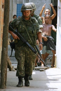 Rio de Janeiro, BRAZIL:  Soldiers patrol a street of the Manguinhos shantytown as resident boys gestures on their backs, 08 March 2006 in Rio de Janeiro, Brazil. Brazilian Army and police forces occupied nine shantytowns in search of ten FAL 7.62mm assault rifles and one 9mm pistol stolen from an army barrack in Rio de Janeiro last Friday.     AFP PHOTO / VANDERLEI ALMEIDA  (Photo credit should read VANDERLEI ALMEIDA/AFP/Getty Images)