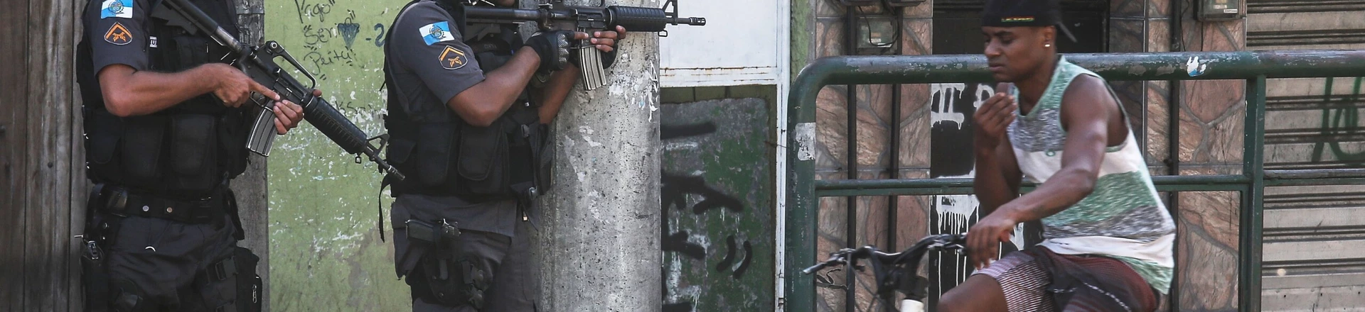RIO DE JANEIRO, BRAZIL - NOVEMBER 20: Military Police (PM) officers hold their weapons as a young man bikes past in the Cidade de Deus 'City of God' favela community during an ongoing police operation on November 20, 2016 in Rio de Janeiro, Brazil. Seven local young men were found killed in the area today following a prolonged firefight between police and suspected gang members. In addition, four police officers were killed yesterday when their support helicopter crashed nearby during the firefight. According to preliminary indications the helicopter was likely not shot down. (Photo by Mario Tama/Getty Images) RIO DE JANEIRO, BRAZIL - NOVEMBER 20: Military Police (PM) officers hold their weapons as a young man bikes past in the Cidade de Deus 'City of God' favela community during an ongoing police operation on November 20, 2016 in Rio de Janeiro, Brazil. Seven local young men were found killed in the area today following a prolonged firefight between police and suspected gang members. In addition, four police officers were killed yesterday when their support helicopter crashed nearby during the firefight. According to preliminary indications the helicopter was likely not shot down. (Photo by Mario Tama/Getty Images)