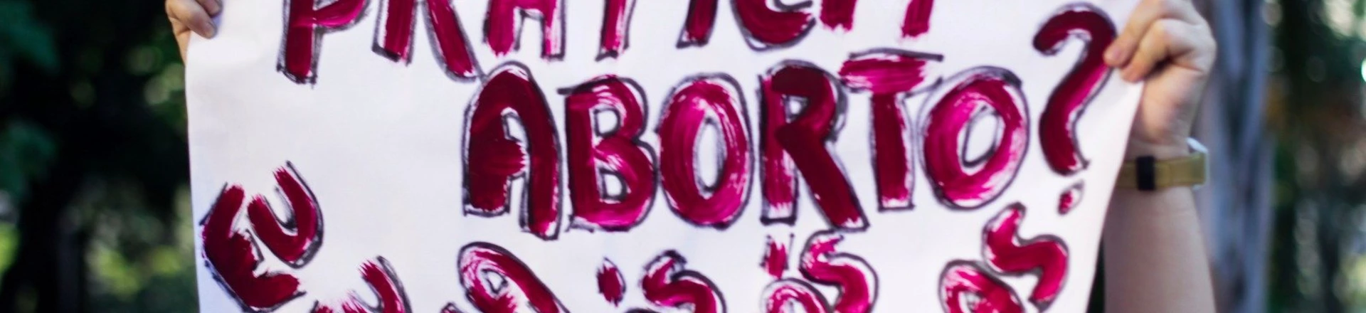 Manifestante com o rosto pintado de Catrina segura cartaz com as palavras "Quem pratica aborto? Eu, tu, ela, nós, vós, elas". Manifestante com o rosto pintado de Catrina segura cartaz com as palavras "Quem pratica aborto? Eu, tu, ela, nós, vós, elas".