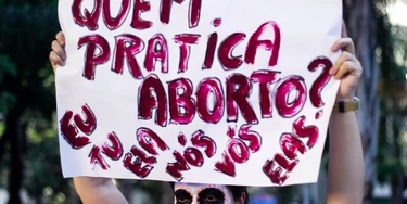 Manifestante com o rosto pintado de Catrina segura cartaz com as palavras "Quem pratica aborto? Eu, tu, ela, nós, vós, elas". Manifestante com o rosto pintado de Catrina segura cartaz com as palavras "Quem pratica aborto? Eu, tu, ela, nós, vós, elas".