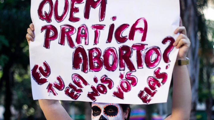 Manifestante com o rosto pintado de Catrina segura cartaz com as palavras "Quem pratica aborto? Eu, tu, ela, nós, vós, elas".